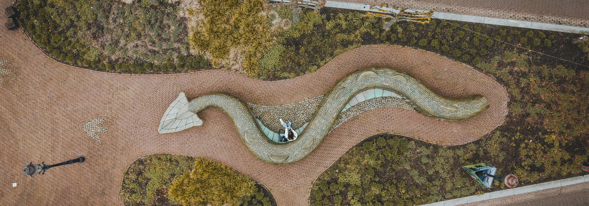Bovenaanzicht van een slingerend kunstwerk in de vorm van een drakenstaart tussen wandelpaden en beplanting.