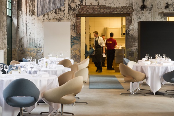 De koks van restaurant One zijn in de achtergrond druk bezig in de keuken terwijl de stoelen en tafels gedekt zijn en klaar staan voor de gasten in een industriële setting