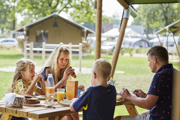 Gezin zit aan een houten picknicktafel en ontbijt in de buitenlucht op camping de Leistert in Roggel, met tenten en groen op de achtergrond.
