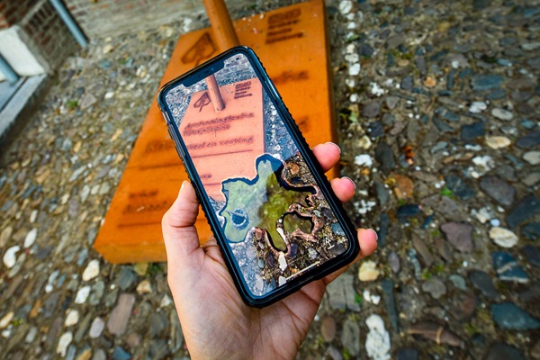 Een hand houdt een smartphone vast, waarop een AR (augmented reality) weergave van een archeologische vondst te zien is. Op de achtergrond staat een roestkleurige speer van de Archeo Route Limburg op een bestrate ondergrond.