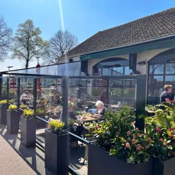 Gasten zitten op het zonnige terras van Gasterij de Knip in Wessem, met uitzicht op de Maas en de jachthaven.