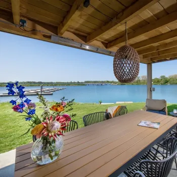 Terras van een vakantiewoning op Parc Maasresidence Thorn, met eettafel, bloemen en vrij uitzicht over het water en de steigers.